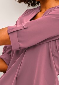 Blusa rosa con una vestibilità rilassata, caratterizzata da manicotti arrotolati, un dettaglio con bottone e un tessuto liscio e leggero. Presente un dettaglio di drappeggio diagonale.