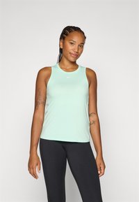 Lachende vrouw met gevlochten haar, gekleed in een mintgroene sporttop en zwarte leggings, staand tegen een effen achtergrond.