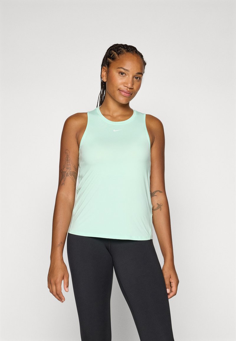 Lachende vrouw met gevlochten haar, gekleed in een mintgroene sporttop en zwarte leggings, staand tegen een effen achtergrond.