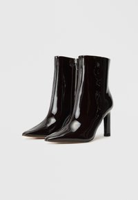 Botas de tornozelo pretas em verniz com bico fino, salto alto e fecho lateral, apresentando um acabamento brilhante e textura suave.