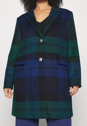 Kobieta w granatowo-zielono-czarnym płaszczu w duże kratki z srebrnymi guzikami, na czarnej bluzce i niebieskich spodniach, zdjęcie przycięte powyżej szyi.