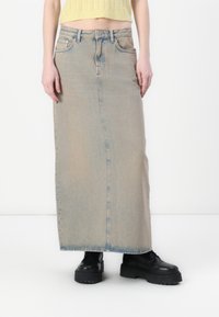 Fustă maxi din denim, de culoare albastru deschis, cu o cusătură frontală, două buzunare laterale și margine destrămată, asortată cu pantofi cu talpă înaltă negri.
