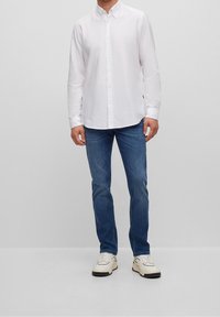 Vitt skjort med knappar framtill, kombinerad med blå jeans och vita sneakers. Skjortan har en klassisk krage och knappslå. Enkel design utan mönster.