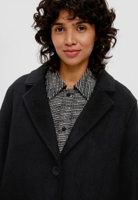 Schwarzer Wollmischmantel mit Reverskragen, mit einem Knopfverschluss, über einem strukturierten schwarz-weißen gestreiften Hemd getragen. Lockige Haare sichtbar.