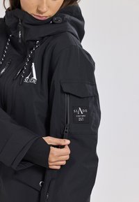 Giacca nera impermeabile con colletto alto, tasche con zip, dettaglio del logo e polsini elastici. Presenta un cappuccio regolabile con cordino e vestibilità adattabile.