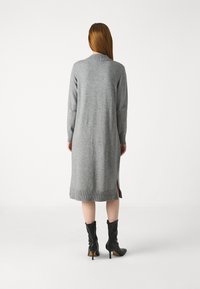 Abito grigio lavorato a maglia con maniche lunghe, spacchi laterali e dettagli a coste sull'orlo, indossato con stivali neri con tacco alto.