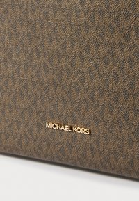 Brūna ādas izskata maku ar logo un teksturētu ģeometrisku rakstu. Zīmola nosaukums "MICHAEL KORS" ir iepresēts zeltā.
