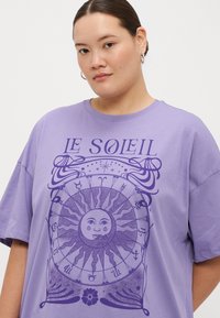 Camiseta oversized morada con un gráfico de sol, símbolos astrológicos y el texto "LE SOLEIL" en morado oscuro. Material de mezcla de algodón suave.