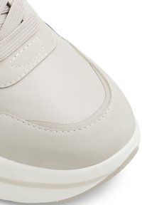 Lys beige sneaker med glat syntetisk overdel, stikninger og rund tå. Har en hvid tekstureret sål og flade snørebånd.