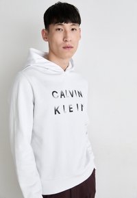 Bela kapuca iz bombaža s sprednjim tiskom, ki bere "CALVIN KLEIN" v gradientno črnih črkah, z vrvico za nastavljanje kapuce in rebrastimi manšetami.