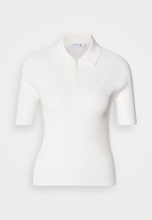 Maglietta bianca a polo, con maniche corte e colletto con zip, caratterizzata da una sottile trama a coste e un piccolo logo Lacoste sul petto.
