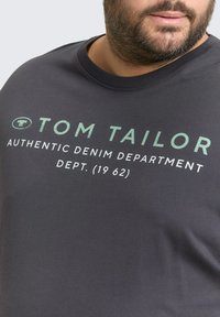 Donkergrijze katoenen t-shirt met een ronde hals. De voorkant toont "TOM TAILOR" en "AUTHENTIC DENIM DEPARTMENT" in groene en witte tekst.