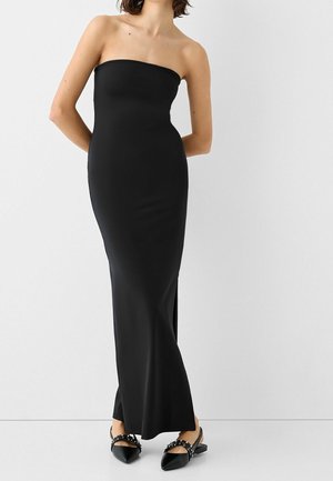 Femme portant une robe noire longue sans bretelles, accompagnée de ballerines noires pointues avec des brides cloutées, debout devant un mur blanc uni.