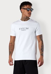 White cotton t-shirt, short sleeves, featuring "ESTEEM MARBELLA" print in black. Paired with black shorts and sunglasses. Smooth texture.
