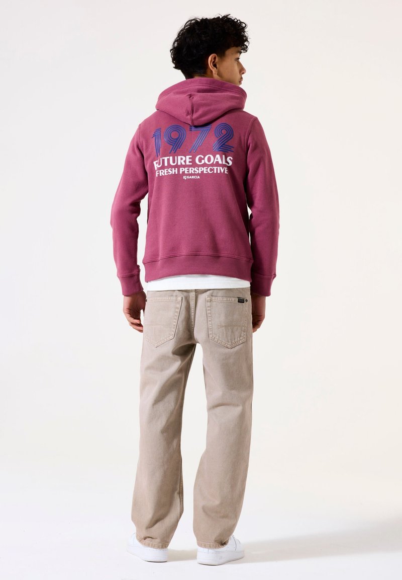 Sudadera color granate con gráficos en blanco y azul que dicen "OBJETIVOS DEL FUTURO PERSPECTIVA FRESCA 1972", combinada con pantalones beige y zapatillas blancas.