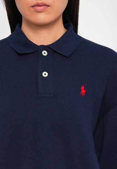 Persoană îmbrăcată cu un tricou polo bleumarin, cu două butoane albe și un mic logo roșu brodat pe partea stângă a pieptului.
