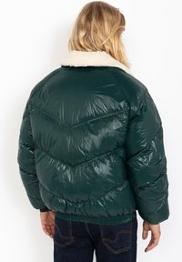 Dark green puffer jacket with a chevron pattern, featuring a beige faux fur collar, shiny texture, and ribbed hem detail at the waist.