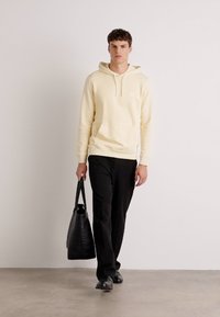 Felpa color crema con tasca frontale, abbinata a pantaloni neri. Modello porta una borsa a tracolla nera e indossa calzature nere.