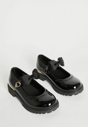 Scarpe in vernice nera con un tacco a blocco basso, punta tonda e fiocchi neri a coste. Presentano un accento dorato e dettagli con margine ondulato.