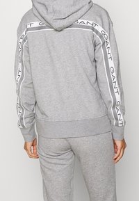 Felpa grigia con cappuccio e maniche lunghe, caratterizzata da un motivo a strisce bianche e dal logo "GANT" ripetuto lungo le spalle. Tessuto con texture morbida.