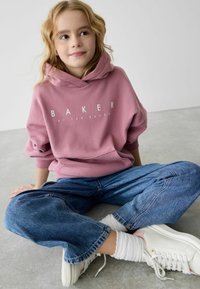 Felpa con cappuccio rosa con stampa "BAKER", abbinata a jeans di denim blu oversize e sneakers bianche con accenti rosa. Tessuto testurizzato visibile.