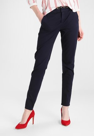 Frau trägt navyblaue Slim-Fit-Hose, ein weißes Hemd mit roten Streifen, einen schwarzen geflochtenen Gürtel und rote Pumps, steht mit einer Hand in der Hosentasche.
