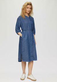 Jeanskleid mit rundem Ausschnitt, drei Viertel Ärmeln, tailliertem Schnitt und Seitentaschen. Es hat ein knielanges Design mit seitlichen Schlitzen.