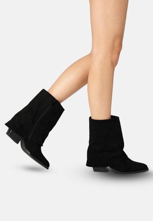 Jambes portant des bottes en daim noir mi-mollet avec des talons blocs bas, un pied relevé et l'autre à plat, présentées sur un fond uni.