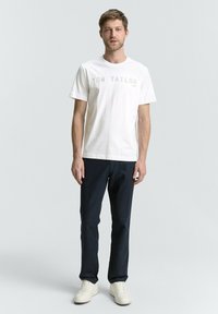 Camiseta de algodón blanca con "TOM TAILOR 1982" impreso en gris claro. Combinada con pantalones azul oscuro y zapatillas blancas. Diseño simple y casual.
