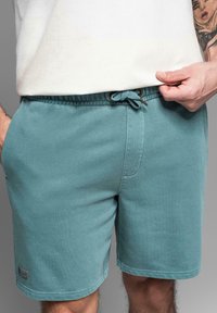 Světlé tyrkysové šortky vyrobené z jemné látky, s elastickým pasem na šňůrku, bočními kapsami a malou logem štítku na lemu.