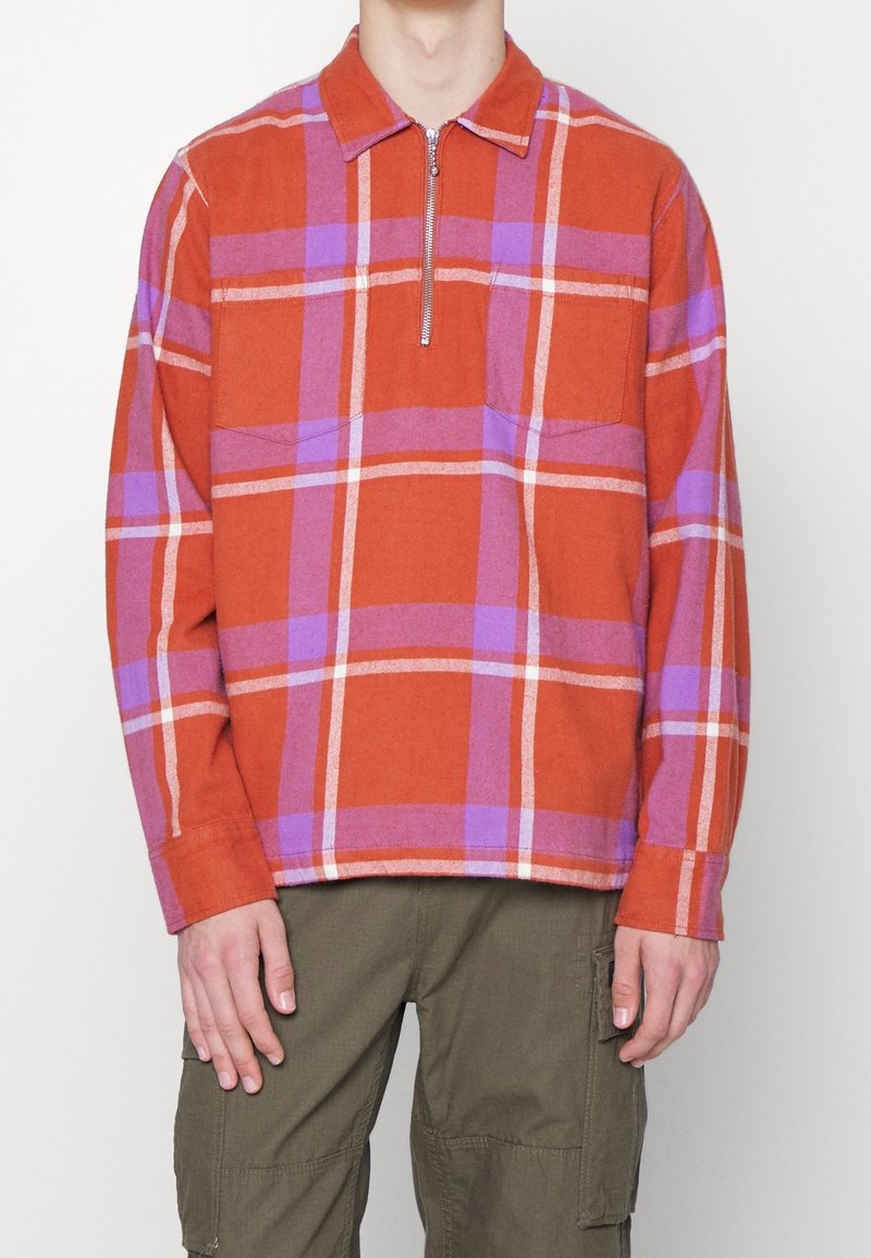 Homme portant une chemise à manches longues à carreaux rouges et violets avec fermeture éclair au col, et un pantalon cargo vert olive, debout devant un fond uni.