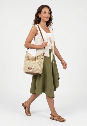 Kvinde går iført en hvid strikket vest, olivengrøn nederdel, brune sandaler og bærer en beige hækle- skuldertaske med en brun rem.