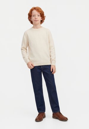 Enfant aux cheveux roux portant un pull beige, un jean bleu foncé et des chaussures marron, debout devant un fond clair et uni.