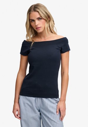 Mujer rubia con top negro sin hombros y pantalones rayados azul claro, de pie frente a un fondo liso.