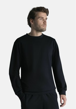 Lyhytukkainen mies, jolla on yksivärinen musta pitkähihaisen paita ja mustat housut, seisoo käsi taskussa ja katsoo oikealle.