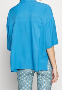 Blauw kortearm shirt van een gladde stof, voorzien van een kraag en een losse snit. Gecombineerd met gestreepte lichtblauwe strakke broek.