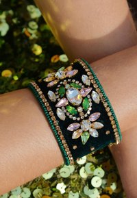 Bracelet en velours vert avec des pierres précieuses multifacettes de différentes formes, entourées de petites perles dorées et de détails complexes.