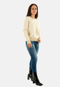 Jerséis crema con un patrón texturizado, combinado con jeans azules y botas de tacón negras. El fondo es blanco liso.