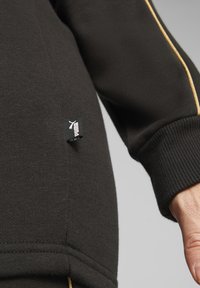 Musthave pusa mustmust detalj sisaldab kuldset aktsenttriipa, ribakätte ja väikest valget logo silti Puma brändimisega. Pehme tekstiilmaterjal.