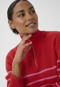 Czerwony sweter dzianinowy z wysokim kołnierzem i złotym zamkiem. Ma różowe poziome paski. Teksturowana tkanina i dopasowany krój.