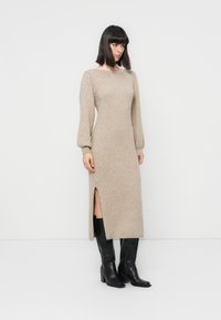 Robe midi en tricot beige avec de longues manches bouffantes, fente sur le côté et encolure ronde, associée à des bottes noires jusqu'aux genoux. Texture lisse.
