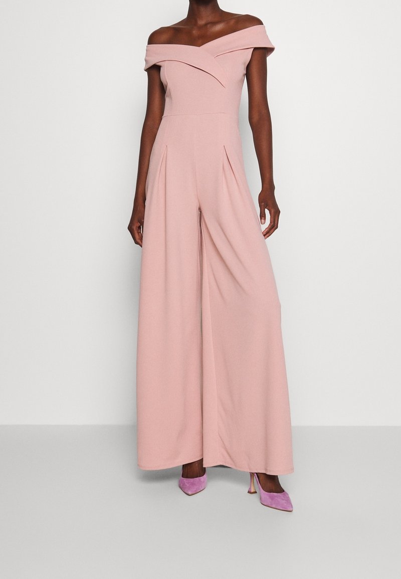 Femme portant une combinaison rose pâle à épaules dénudées avec jambes larges, associée à des chaussures à talons hauts violettes, debout devant un fond uni.