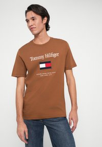 Rjava bombažna majica s kratkimi rokavi, ki ima belo "Tommy Hilfiger" logotip ter pisan našitek z rdečimi, belimi in modrimi pravokoti na prsih.