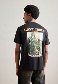Schwarzes Baumwoll-T-Shirt mit einem Rückenmuster von Kakteen, dem Text "CAN'T TOUCH THIS" in Orange und "A LITTLE BIT PRICKLY" in Weiß.