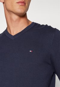 Pull en tricot bleu marine à col en V avec texture côtelée et petit logo brodé en rouge, blanc et bleu sur la poitrine gauche.