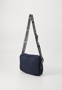 Námorná crossbody taška s textúrovaným materiálom, obdĺžnikovým tvarom, zipsovým vreckom a čiernym popruhom s bielym nápisom "Nørgaard".