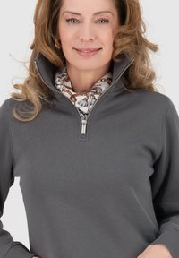 Felpa grigia con zip e colletto alto, con una sciarpa bianca a fantasia sotto. Texture liscia e design casual. Dettaglio della zip in argento.