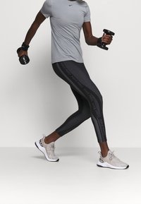 T-shirt de sport gris, leggings noirs à motifs avec le logo "NIKE PRO" et baskets gris clair. Personne tenant des haltères dans une pose dynamique.