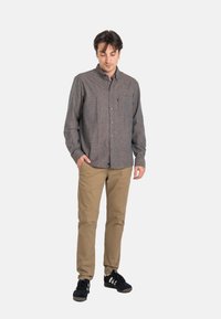 Hombre de pie con la cabeza ligeramente inclinada hacia abajo, vestido con una camisa gris abotonada, pantalones beige y zapatillas negras, frente a un fondo blanco liso.