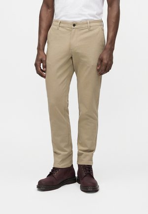 Mann trägt beige gerade geschnittene Hose und dunkelbraune Schnürstiefel und steht vor weißem Hintergrund.